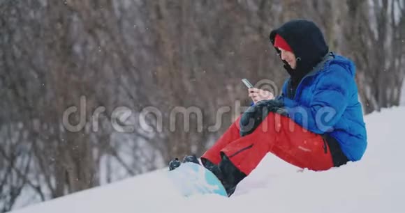 一位坐在雪地上的男性滑雪者通过电话为社交网络拍摄一处美丽的度假胜地景观视频的预览图