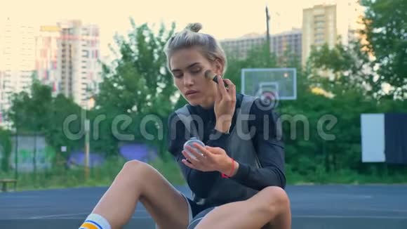 漂亮的女篮球运动员坐在球场上做化妆公园和背景建筑视频的预览图