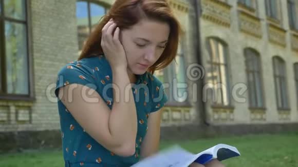可爱的生姜学生看书对着镜头微笑坐在大学附近的公园里在背景下建筑快乐视频的预览图