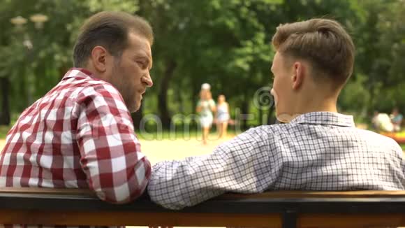胡子爸爸和十几岁的儿子交流坐在公园的长凳上信任视频的预览图