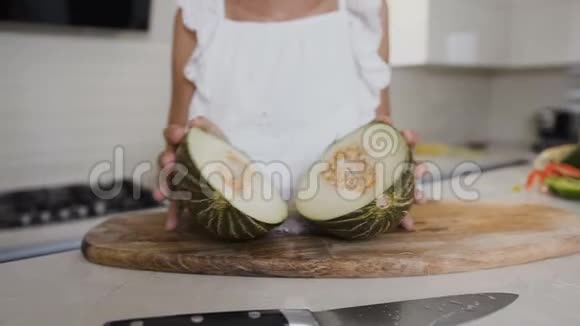 这位穿着白色连衣裙的可爱少女手中拿着一个切成两半的甜瓜看着家厨房里的相机关闭视频的预览图