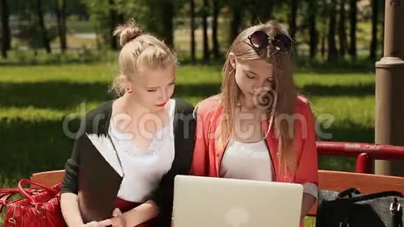两个年轻漂亮的女学生手拿笔记本电脑在绿色公园的长凳上学习前景视频的预览图