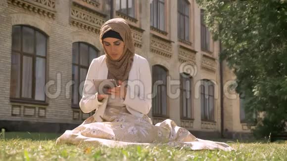 美丽的穆斯林妇女戴着头巾在平板电脑上打字坐在大楼附近的公园的草地上迷人的女性带着小玩意视频的预览图