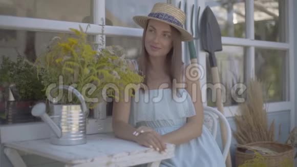 美丽的年轻女子戴着草帽穿着白色的连衣裙坐在小村庄的房子前面农村生活方式慢慢视频的预览图