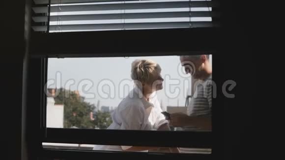 幸福微笑的浪漫情侣站在阳光明媚的小阳台上透过公寓窗户观看使用智能手机视频的预览图