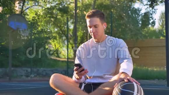 帅气快乐的年轻人坐在篮球场上打电话抱着球听音乐微笑着视频的预览图