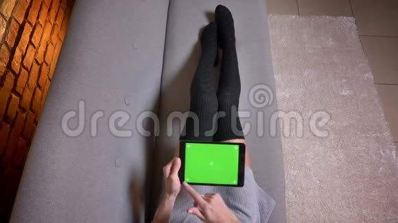 室内沙发上穿着可爱袜子的女腿特写镜头女人水平地拿着绿色屏幕的平板电脑视频的预览图