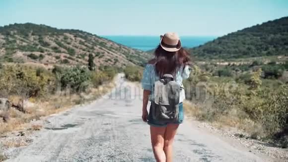 后景活跃的背包客女人戴着帽子走在被群山环绕的海上小路上视频的预览图