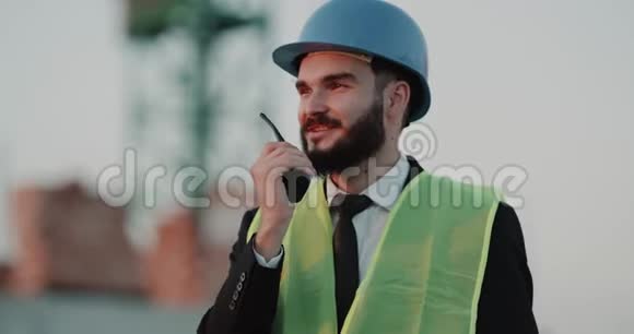 施工现场的年轻工程师戴着安全帽用收音机与工人交谈视频的预览图