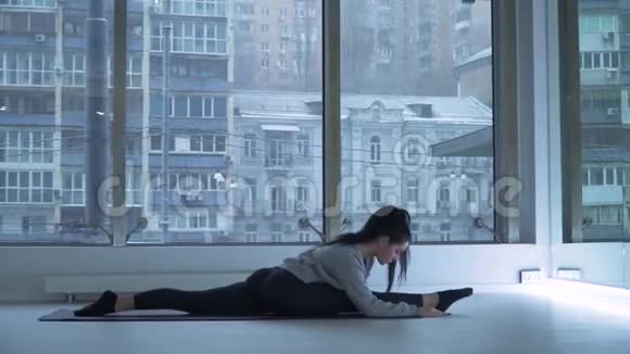 年轻灵活的女人在舞厅里的垫子上伸展双腿大的落地窗美丽的城市景观视频的预览图