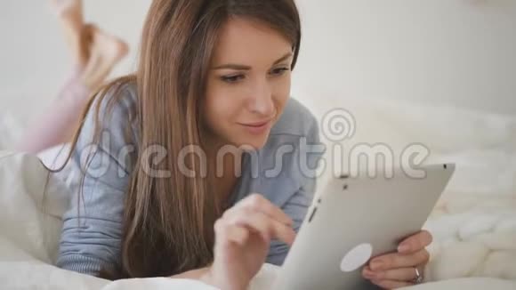 穿着蓝色衬衫棕色头发的女人躺在床上铺着白色床单上网冲浪视频的预览图