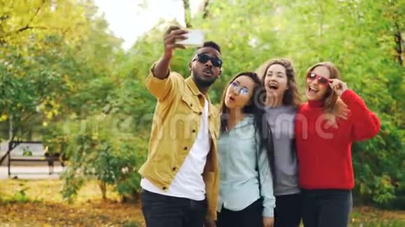 非洲裔美国学生正在与美丽的女孩自拍亚洲和高加索人站在公园里使用智能手机视频的预览图