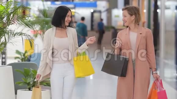 两个快乐的年轻女人慢吞吞地拎着销售袋穿过购物中心视频的预览图