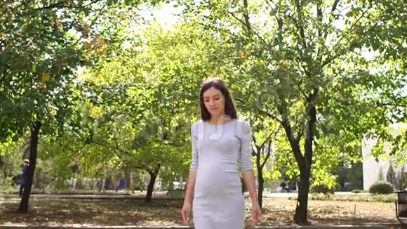 快乐的白种人怀孕女孩抚摸她的腹部站在公园的大树附近视频的预览图
