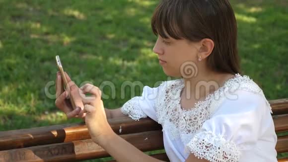 美丽的女孩使用智能手机正在一个美丽的绿色公园的长凳上写一封信慢动作青年视频的预览图