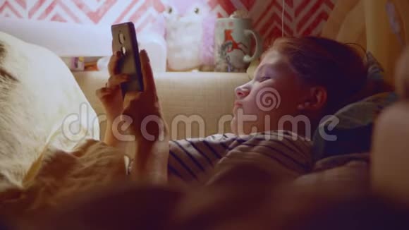 年轻的金发女孩躺在床上手里拿着智能手机睡觉前上网视频的预览图