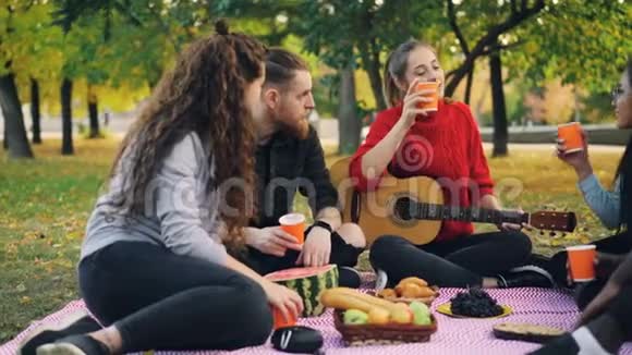 在温暖的秋日年轻的男人和女人在公园野餐时用吉他品尝和敲击眼镜友谊视频的预览图