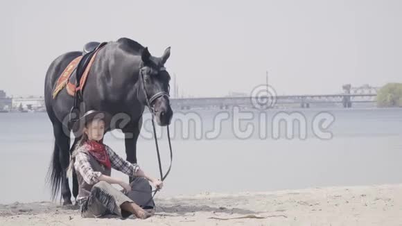 穿牛仔衣服的女人和她的马坐在沙滩上4K视频的预览图