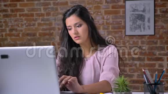 年轻认真的女孩在阁楼办公室工作手提电脑打字看着相机微笑背景砖墙视频的预览图