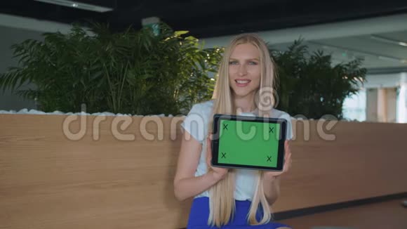 在办公室展示平板电脑的女人年轻的金发女郎带着绿色屏幕的平板电脑看着摄像机视频的预览图