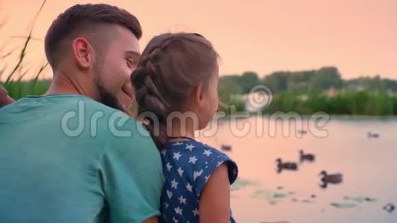 英俊的父亲和他的小女儿坐在湖边看鸭子聊天日落家庭观念视频的预览图