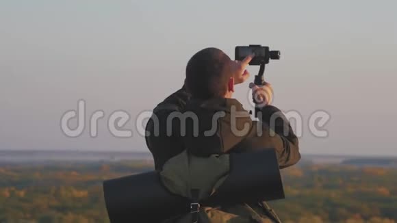 这位游客拿着稳定器相机在日落背景上有一部手机他自拍拍视频视频的预览图