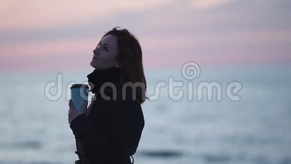 年轻女子在海滩上喝着咖啡在寒冷的蓝色夕阳下清澈的倒影在水面上视频的预览图