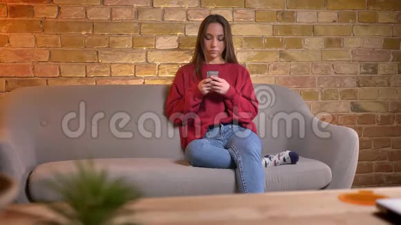 年轻的白种人黑发女孩在智能手机上打字的肖像坐在家里的沙发上视频的预览图
