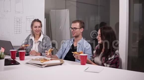 年轻的白种人同事午休时一起在办公室吃披萨笑说话玩得开心工作人员视频的预览图