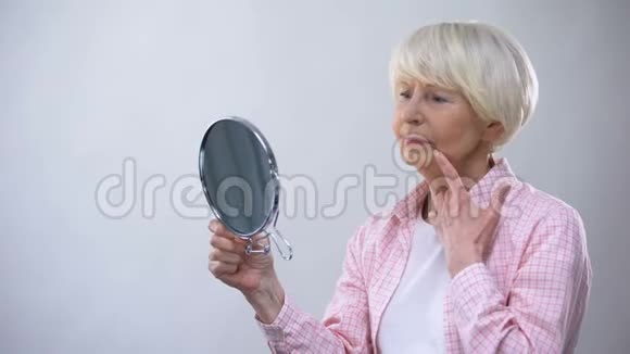 一位老年妇女看着镜子里布满皱纹的脸想着整容手术视频的预览图