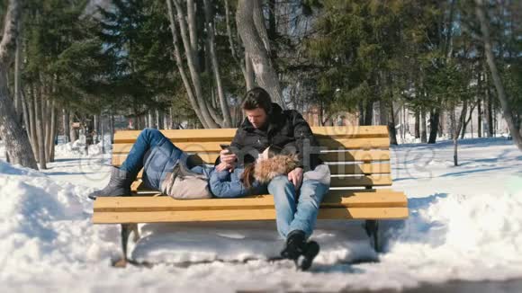 男人和女人一起在冬季城市公园的长凳上休息冬日阳光明媚在手机上看一些东西视频的预览图