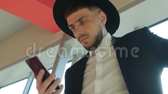 穿着西装戴着一顶带胡须的帽子的时尚白种人男性商人站在智能手机上阅读信息视频的预览图