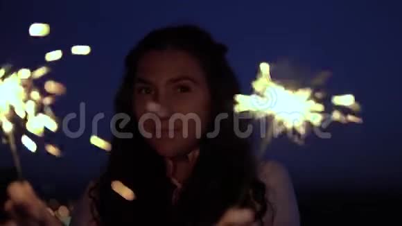 一个留着深色长发的年轻女孩站在夜幕降临的城市背景下手里拿着烟火慢动作视频的预览图