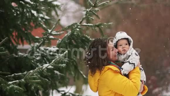 可爱的年轻女子穿着黄色羽绒服和婴儿穿着工作服走路下雪了妈妈给他的儿子看视频的预览图