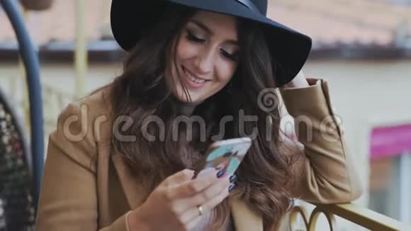 穿着一顶黑色经典帽子和一件米色外套的年轻黑发特写甜美地微笑着在她的智能手机读短信慢慢视频的预览图