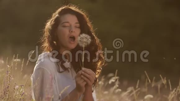 美丽的年轻女子在夏日夕阳下的麦田里吹蒲公英美丽与夏日概念视频的预览图
