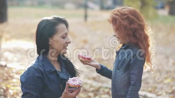 一个漂亮的妈妈和她漂亮的红头发的女儿站在一个秋天的公园里他们吃甜甜圈互相喂吃视频的预览图