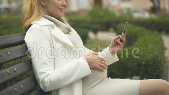 快乐的孕妇滚动智能手机照片在长凳上的公园放松视频的预览图