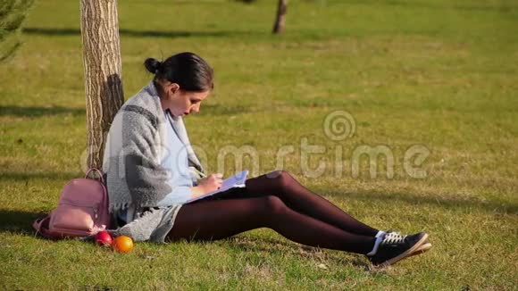 穿着短裤和运动鞋的漂亮长腿可爱的黑发女孩坐在公园附近的草地上在笔记本上写字视频的预览图
