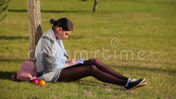 穿着短裤和运动鞋的漂亮长腿可爱的黑发女孩坐在公园附近的草地上在笔记本上写字视频的预览图