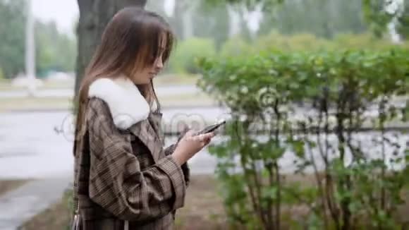 白天下雨后独自一个黑发女孩走在城市的秋日潮湿的街道上在智能手机上打字视频的预览图
