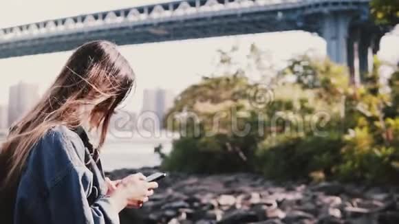 年轻的旅行女孩带着背包在平静的布鲁克林大桥河岸边散步带智能手机慢动作视频的预览图