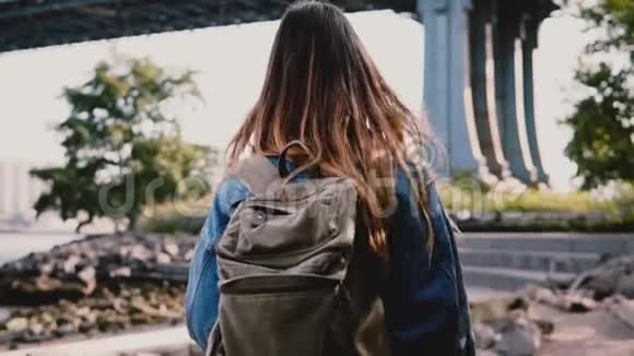 镜头跟随年轻的旅行者女孩背着背包沿着美丽的阳光明媚的布鲁克林公园河岸慢动作视频的预览图