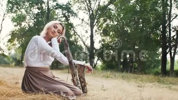 年轻漂亮的浪漫女孩坐在地上的干草上手里拿着萨克斯管一个拿着乐器的女人视频的预览图
