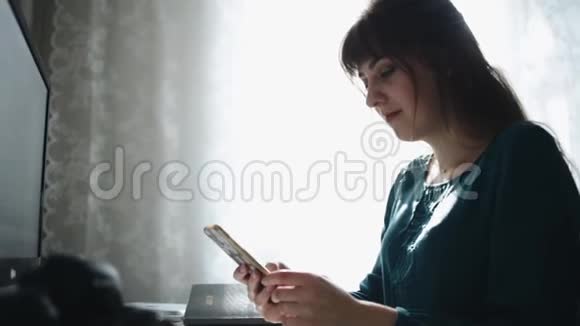 一个漂亮女孩坐在笔记本电脑前在她的智能手机上打字时的近景视频的预览图