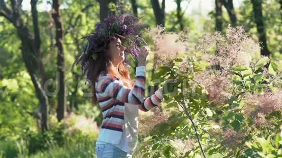 一个美丽的女人头上戴着一朵矢车菊在森林里的灌木丛中她的头发粉红色的花朵视频的预览图