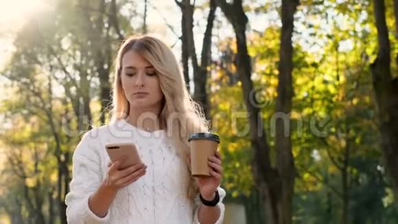 时髦的年轻可爱的微笑女孩使用现代智能手机同时在城市公园散步女士输入短信视频的预览图