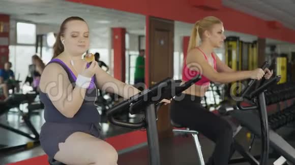 快乐的超重女士在健身房骑运动自行车时吃面包视频的预览图