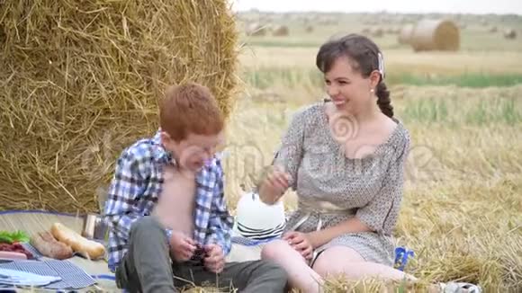夏夜一位美丽的年轻女子带着红头发的儿子在麦田中央的干草堆附近野餐视频的预览图