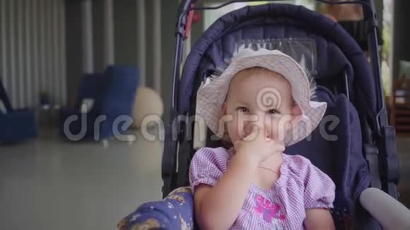 戴帽子的小女孩坐在蓝色的婴儿车里用手舔嘴动作缓慢视频的预览图
